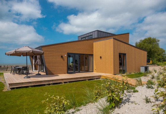 Moderne lodge med træbeklædning, stor terrasse og have under en blå himmel med spredte skyer.