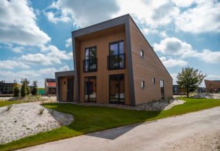 Moderne lodge met schuin dak, grote ramen en balkon, omgeven door groen gras en blauwe lucht.
