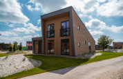 Modern lodge with a slanted roof, large windows, and balcony, surrounded by green grass and a blue sky.