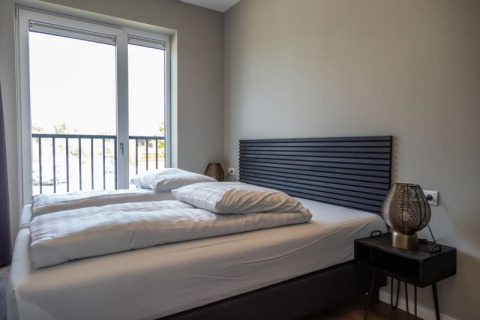 Chambre moderne dans un lodge avec lit double, grande fenêtre, tête de lit grise et table de nuit.