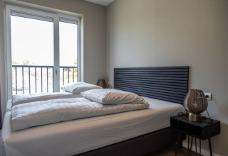 Chambre moderne dans un lodge avec lit double, grande fenêtre, tête de lit grise et table de nuit.