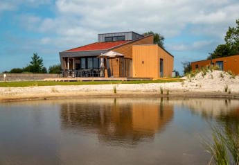 Moderne lodge aan een vijver met terras, brede ramen en parasol, omzoomd door graslanden en natuur.