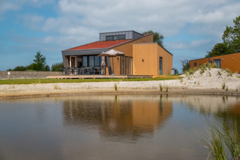 Lodge moderne au bord d’un lac avec terrasse en bois, grandes baies vitrées et parasol sous un ciel clair.