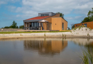 Lodge moderne au bord d’un lac avec terrasse en bois, grandes baies vitrées et parasol sous un ciel clair.