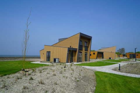 Lodges modernes aux toits inclinés, pelouses vertes et environnement côtier sous un ciel bleu éclatant.