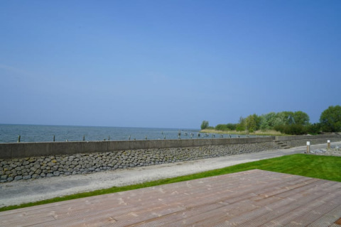 Blick von einer Lodge auf das Wasser mit Steinmauer, grünem Rasen und Holzterrasse bei blauem Himmel.