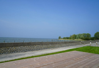 Vue d’une lodge sur l’eau avec un mur en pierre, pelouse verte et terrasse en bois sous ciel bleu.