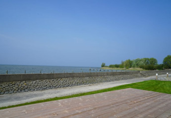 Blick von einer Lodge auf das Wasser mit Steinmauer, grünem Rasen und Holzterrasse bei blauem Himmel.