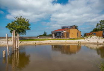 Et moderne lodgehus med terrasse ved en lille sø, omgivet af sand og grønne træer under en blå himmel.