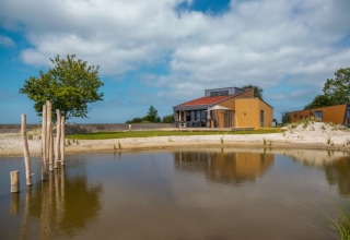 Un albergue moderno con terraza junto a un pequeño estanque, rodeado de arena y árboles bajo cielo azul.