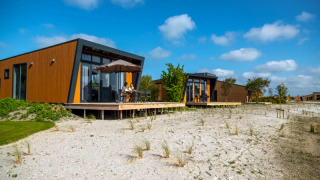 Moderne lodges met houten gevels en grote ramen op een zandperceel onder een blauwe, zonnige lucht.