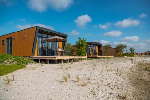 Cabañas modernas con fachadas de madera y ventanas grandes sobre suelo arenoso bajo un cielo azul soleado.