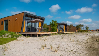 Cabañas modernas con fachadas de madera y ventanas grandes sobre suelo arenoso bajo un cielo azul soleado.
