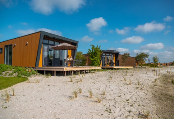 Modern lodges with wooden facades and large windows on a sandy lot under a blue, sunny sky.