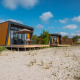 Cabañas modernas con fachadas de madera y ventanas grandes sobre suelo arenoso bajo un cielo azul soleado.