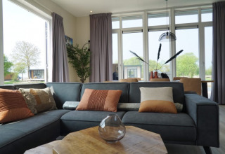 Salón de lodge moderno con grandes ventanas, sofá gris, cojines decorativos y vistas al exterior verde.