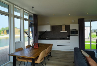 Lodge moderne avec cuisine ouverte, table en bois et grandes fenêtres donnant sur l’eau et la nature.