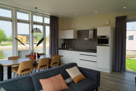 Moderne lodge met open keuken, eettafel voor zes, bank en grote ramen met uitzicht op water en natuur.