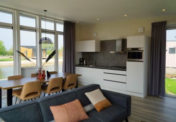 Moderne lodge med åbent køkken, spisebord til seks og sofa, store vinduer med udsigt til natur og vand.