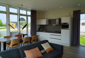 Moderne lodge med åbent køkken, spisebord til seks og sofa, store vinduer med udsigt til natur og vand.