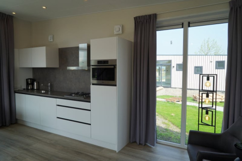 Intérieur de lodge moderne avec cuisine blanche, grande baie vitrée, rideaux gris et vue sur jardin.