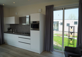 Intérieur de lodge moderne avec cuisine blanche, grande baie vitrée, rideaux gris et vue sur jardin.