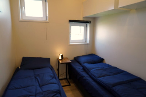 Kleine lodgekamer met twee eenpersoonsbedden, blauwe dekbedden en een tafeltje met lamp ertussen.