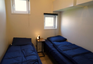 Kleine lodgekamer met twee eenpersoonsbedden, blauwe dekbedden en een tafeltje met lamp ertussen.
