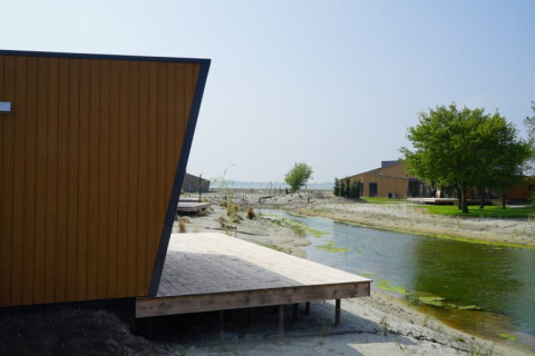 Lodge moderne près d’un canal étroit avec terrasse en bois, sol sableux et arbres verts sous ciel bleu.