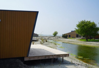 Lodge moderne près d’un canal étroit avec terrasse en bois, sol sableux et arbres verts sous ciel bleu.