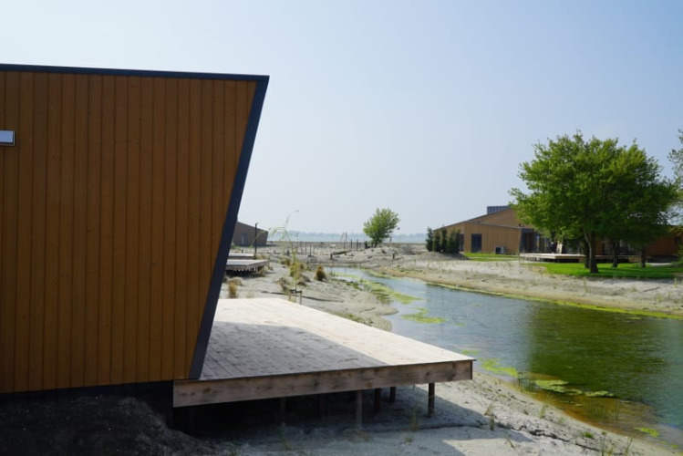 Lodge moderno junto a un canal estrecho, terraza de madera, suelo arenoso y árboles verdes.