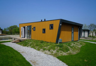 Un lodge moderne en bois aux grandes fenêtres, posé sur une pelouse verte avec un chemin de gravier ensoleillé.