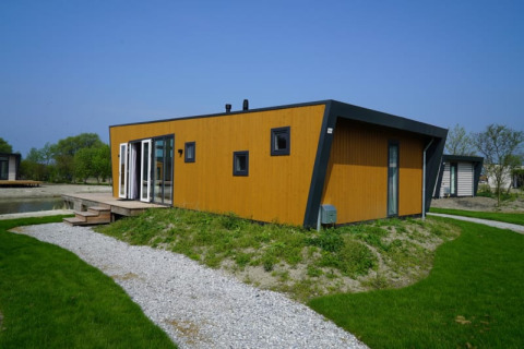 A modern wooden lodge with large windows, situated on green grass, featuring a gravel path and blue sky.