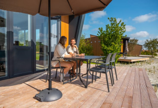 Dos personas conversan sentadas en una terraza de madera frente a una moderna cabaña con sombrilla grande.
