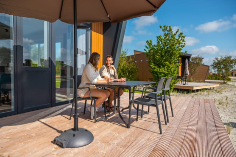 Two people sit and talk at a table on the wooden deck of a modern lodge, shaded by a large umbrella.