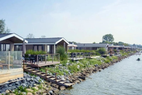 Row of modern lakeside lodges with wooden decks, outdoor seating, lush greenery, and calm water view.