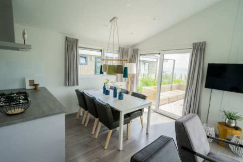 Modern lodge dining area with six chairs, large windows, gray curtains, and a view to a garden patio.