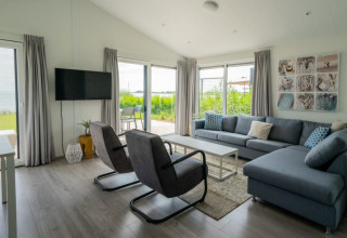 Moderne living in een lodge met grote ramen, grijze zetel, stoelen, tv en uitzicht op de tuin buiten.