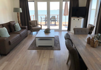 Lyst opholdsområde i en lodge med moderne møbler og store vinduer, der vender ud mod vandet og en terrasse.