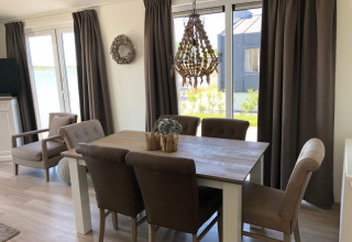 Comedor luminoso en una cabaña con cortinas marrones, mesa de madera, sillas acolchadas y vista al jardín.