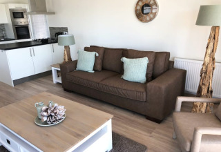 Moderne lodge woonruimte met bruine zetel, mintgroene kussens, boomstamlampen en open keuken achteraan.