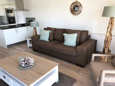 Moderne lodge-stue med brun sofa, lysegrønne puder, trælamper, sofabord og åbent køkken i baggrunden.