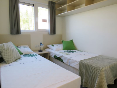 Bright lodge room with two single beds, green pillows, leafy bedding, shelving, and windows with curtains.