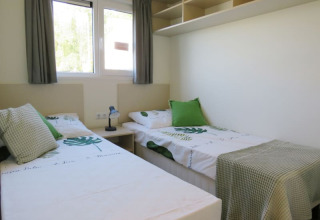 Bright lodge room with two single beds, green pillows, leafy bedding, shelving, and windows with curtains.