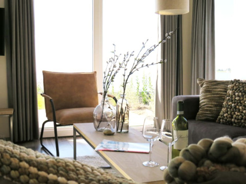 Modernes Wohnzimmer einer Lodge mit großen Fenstern, braunem Stuhl, Dekokissen und Wein auf dem Tisch.