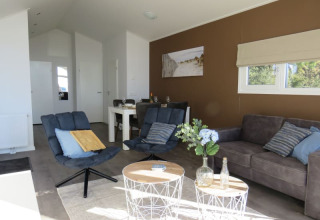 Lodge woonkamer met moderne blauwe stoelen, grijze bank, witte tafels en veel daglicht door ramen.
