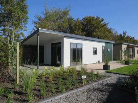 Moderne lodge met terras en tuin in een vakantiepark, omgeven door bomen en een heldere blauwe lucht.