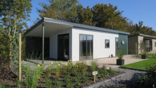 Cabaña moderna con terraza y jardín en un complejo vacacional, rodeada de árboles y cielo azul claro.