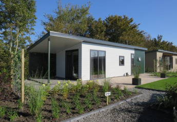 Cabaña moderna con terraza y jardín en un complejo vacacional, rodeada de árboles y cielo azul claro.