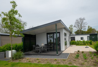 Lodge moderne avec terrasse couverte, coin repas extérieur et jardin, entouré de verdure et d’arbres.